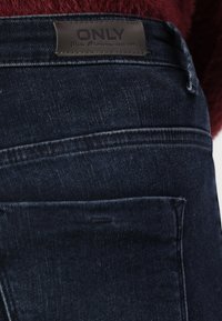 Jeans din denim albastru închis cu o etichetă din piele cu inscriptionarea "ONLY" la talie. Prezintă cusături clasice și buzunare pe spate.