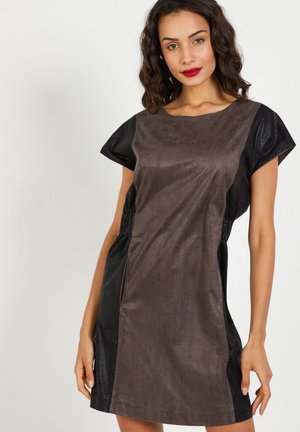 Mujer con cabello oscuro y ondulado, vestida con un vestido corto de mangas cortas en marrón y negro, de pie contra un fondo claro y liso.