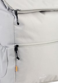 Sac à dos gris présentant un extérieur en textile robuste, de multiples compartiments zippés et une étiquette de logo. Surface lisse avec un motif légèrement texturé.