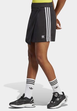 Persoon draagt een zwarte Adidas skort met witte strepen, gecombineerd met witte Adidas sokken en zwarte Adidas sneakers, staand tegen een witte achtergrond.