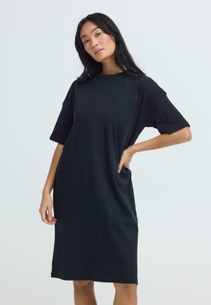 Femme aux longs cheveux noirs portant une robe noire ample arrivant aux genoux, debout avec une main sur la hanche devant un fond clair uni.