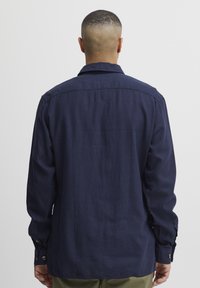 Camisa de manga larga azul marino en tejido ligero, con collar tipo botón, yugo trasero, y puños abotonados para un cierre seguro.