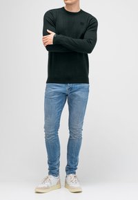 Dunkelgrüner Strickpullover mit Rundhalsausschnitt, kombiniert mit hellblauen Skinny-Jeans und cremefarbenen Sneakers. Dezentes Logo-Detail auf dem Pullover.