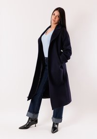 Donna in piedi con un cappotto blu navy, maglione azzurro chiaro, jeans scuri con risvolto e stivaletti neri con punta, su sfondo bianco.