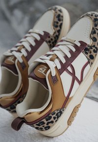 Des baskets avec une tige beige, des accents marron et bourgogne, et un détail en imprimé léopard. Elles présentent des lacets blancs et des semelles texturées.
