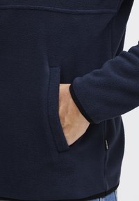 Tamno plavi flis pulover s mekom teksturom, sadrži prednji džep, kontrastne crne detalje na manžetama i diskretan logo detalj sa strane.