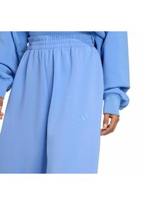 Persona vestida con pantalones de chándal elásticos de cintura alta en azul claro y sudadera a juego, con una mano visible a un lado, pequeño logo en el muslo izquierdo.