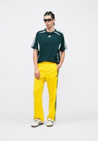 Zielona sportowa koszulka z białymi akcentami, w połączeniu z żółtymi spodniami dresowymi z bocznymi paskami. Obuwie kremowe z ciemnymi detalami.