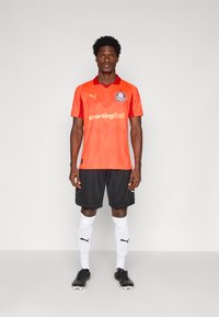 Oranžna nogometna majica s kratkimi rokavi z rdečimi akcenti, okroglo ovratnico in blagovno znamko. Usklajena z črnimi kratkimi hlačami, belimi nogavicami in črnimi kramli.