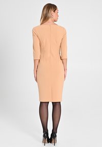 Figurbetontes, knielanges Kleid aus hellbeigem Material mit Dreiviertelärmeln und einem Reißverschluss hinten. Getragen mit schwarzen High Heels.