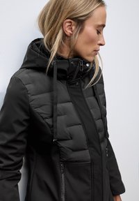 Black jacket featuring a mix of smooth fabric and quilted sections, a hood, zippered pockets, and adjustable drawstrings.