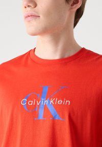 Červené bavlnené tričko so zaobleným výstrihom, krátkymi rukávmi a grafickým potlačou "Calvin Klein" v bielej a modrej farbe na hrudi.