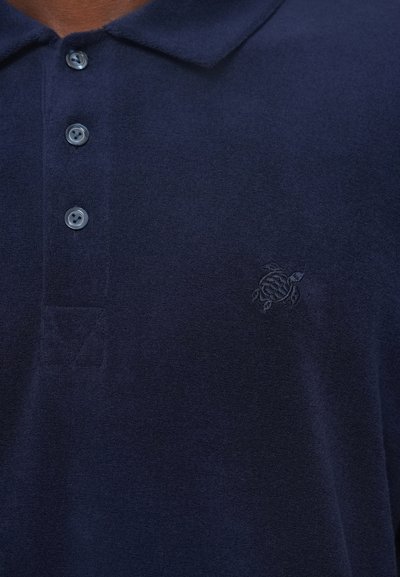 Tums zila polo krekls, izgatavots no mīksta auduma, ar apkakli un piecām pogām. Izšūts bruņurupuču logo kreisajā krūtī.