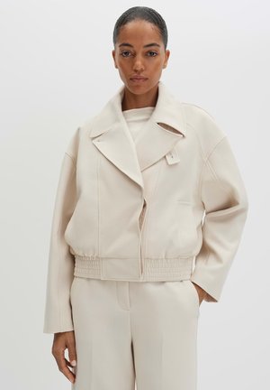 Femme portant une veste oversize beige clair avec un large col et une taille élastique, associée à un pantalon beige assorti, debout devant un fond blanc.