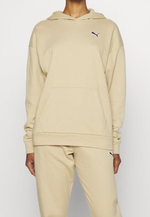 Sweat à capuche beige et pantalon de jogging assorti avec logos Puma noirs sur le torse et la cuisse, porté par une personne debout.
