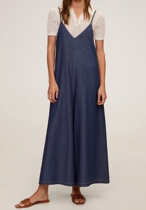 Vrouw draagt een wijde donkere denim jumpsuit over een wit, kort mouwtextuur shirt met bruine platte sandalen, staand tegen een effen achtergrond.