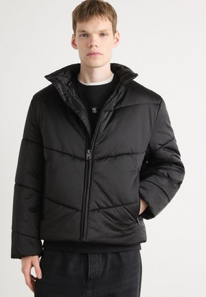 Black puffer jacket with a high collar, quilted design, zip closure, and elasticized cuffs. Worn over a dark sweater with a logo.