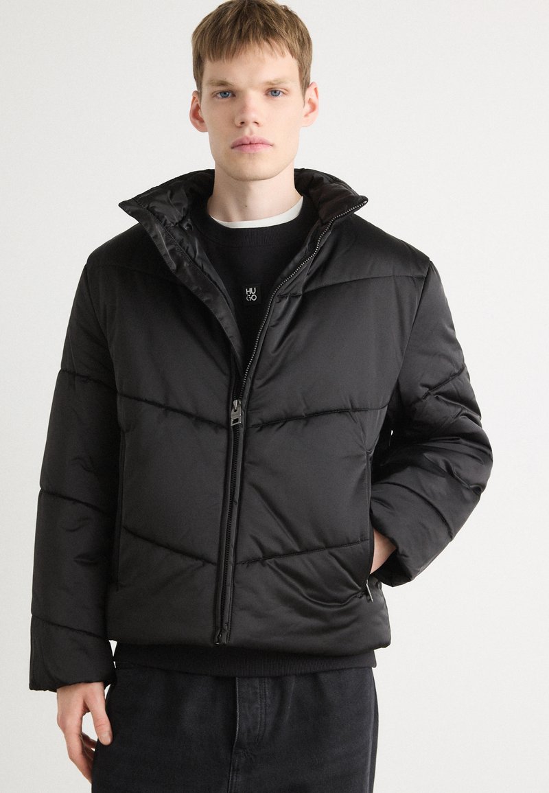 Black puffer jacket with a high collar, quilted design, zip closure, and elasticized cuffs. Worn over a dark sweater with a logo.