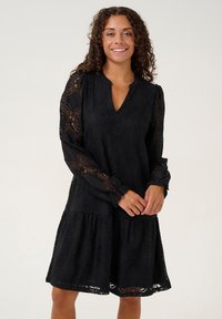 Vestido de encaje negro con mangas largas y transparentes, escote en V y falda en capas con patrones florales. La tela texturizada añade profundidad.