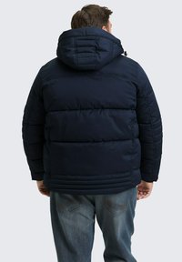 Marineblauwe gewatteerde jas met een capuchon, voorzien van gestikte secties en een elastische zoom. Draagt in combinatie met blauwe jeans, wat een casual uitstraling geeft.