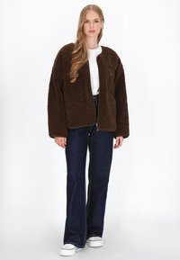 Brown sherpa jacket with a zippered chest pocket, loose fit, and wide sleeves, paired with dark blue jeans and white sneakers.