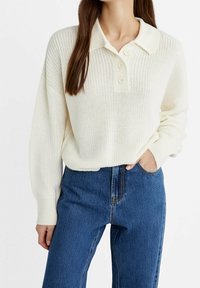 Roomkleurige gebreide trui met een kraag en drie knopen. Gecombineerd met high-waisted blauwe jeans met een klassieke denimtextuur.