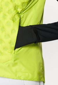 Γιλέκο με επένδυση σε φωτεινό πράσινο λάιμ, με υφή από ανυψωμένες τελείες, τσέπη με φερμουάρ και αντίθετο μαύρο μακρυμάν ικο από κάτω.
