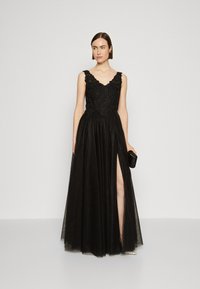 Rochie de seară neagră cu decolteu în V, broderie complexă pe corset, fustă fluidă și slit până la coapsă; asociată cu o poșetă neagră.