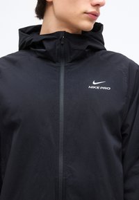 Personne portant une veste zippée Nike Pro noire avec capuche, affichant le logo sur la poitrine gauche, regardant vers la droite.