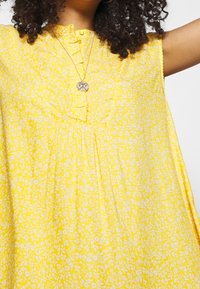 Gul ærmeløs kjole med blomsterprint, med knaplukning ved halsudskæringen og folder, der skaber en løs silhuet.