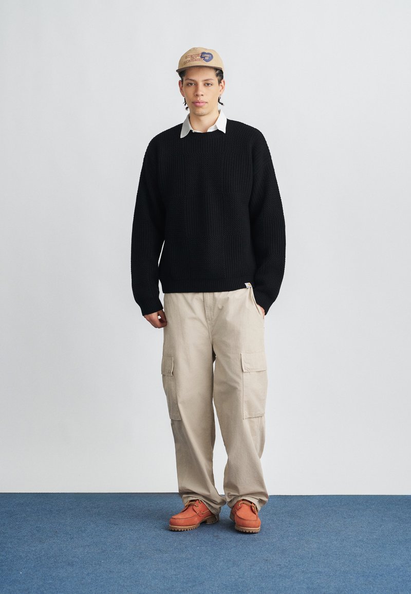 Sort strikket sweater med afslappet pasform, beige cargobukser med flere lommer og orange sko med tyk sål. Beige kasket med logo.