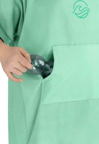 Mintgrünes Kleidungsstück mit kurzen Ärmeln und einer großen Fronttasche. Eine Hand steckt eine klare Plastikflasche in die Tasche.