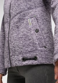 Lila mönstrad jacka med dragkedja, sidofickor och justerbar nederkant. Har varumärkestagg och svarta hårdvarudetaljer. Mjuk textur.