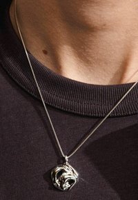Silversmycke i form av ett hängsmycke med en abstrakt, organisk form på ett fint kedja, vilande mot en mörk tygbakgrund.