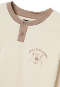 Crème-kleurige t-shirt met een bruine geribbelde kraag, twee knopen en een geprinte cartoonbeer met de tekst "GOEDE OCHTEND!" in lichtbruin.