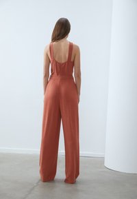 Rustfarget jumpsuit med brede ben, store stropper og lav rygg. Stoffet har en strukturert finish, og det har en belted midje for definisjon.