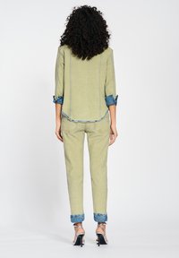Person with curly hair wearing rolled-up sleeves and pants with denim cuffs, standing in silver high-heeled sandals against white backdrop.