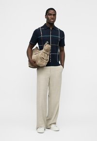 Homme portant un polo bleu marine avec des lignes de grille multicolores, un pantalon beige à jambes larges, des baskets blanches, tenant un sac en daim beige sur fond blanc.