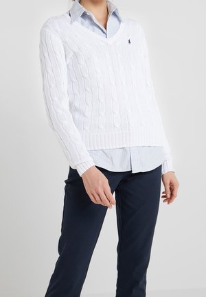 Person, die einen weißen Kabelstrickpullover mit V-Ausschnitt über einem blau-weiß gestreiften Hemd und dunklen Hosen vor einfachem Hintergrund trägt.