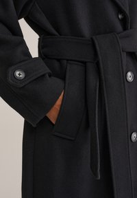Cappotto nero con una morbida texture, caratterizzato da una vita con cintura, dettagli con bottoni e una tasca visibile con foderatura marrone.