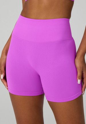 Woman wearing high-waisted, bright purple seamless athletic shorts, showing midriff and hands resting at sides against a gray background.