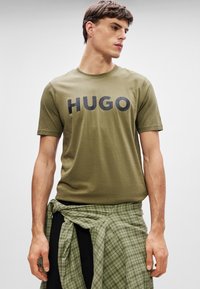 Olivovo zelené bavlnené tričko s okrúhlym výstrihom a krátkymi rukávmi, na prednej strane s výrazným čiernym nápisom "HUGO".