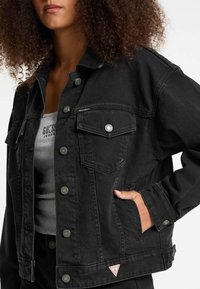 Black denim jacket with button closures, two front pockets, and a small logo badge. Features a classic collar and a relaxed fit design.