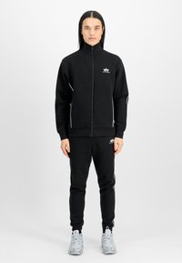 Svart zip-up hettegenser og matchende joggebukser laget av et mykt materiale, med hvite side-striper og en liten logo på brystet.