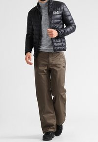 Zwarte pufferjas met horizontaal doorgestikt patroon, grijze coltrui, bruine cargobroek en zwarte schoenen. Eenvoudig ontwerp zonder zichtbare logo's.