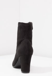 Bottine en daim noir avec un talon haut, une fermeture éclair latérale et une texture lisse. Le talon a une forme arrondie et une base solide.