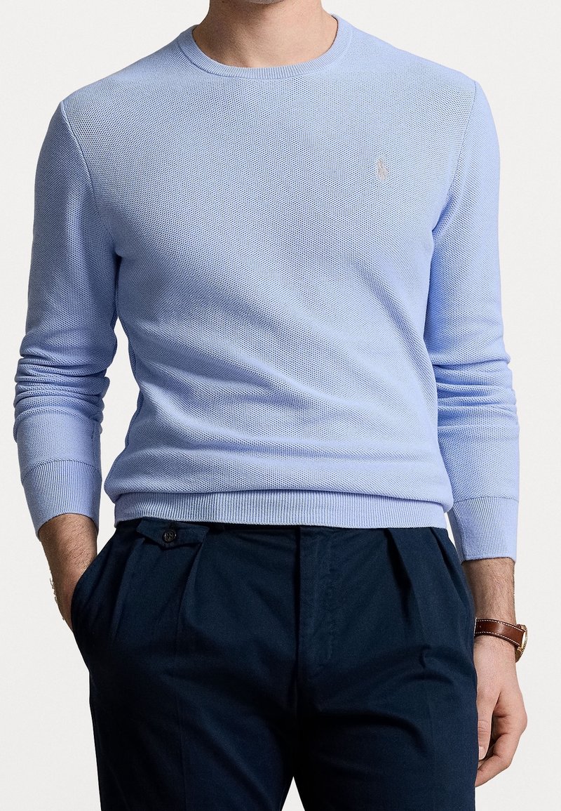 Světlounce modrý pletený svetr s kulatým výstřihem, texturovanou látkou a žebrovanými manžetami, doplněný tmavými kalhotami. Jemný detail loga na hrudi.
