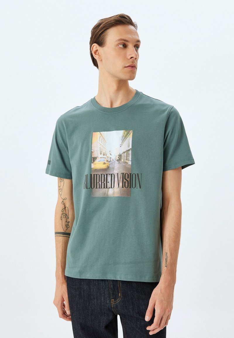 Camiseta de algodón verde con cuello redondo, que presenta un gráfico central de imágenes borrosas de la ciudad y el texto "VISIÓN BORROSA" en una fuente en negrita.