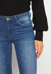 Jeans din denim albastru cu talie înaltă și croială ajustată. Prezintă un închidere cu nasture auriu, buzunare frontale și un model ușor de decolorare.