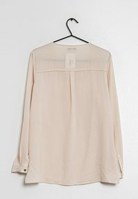 Ljust beige långärmad blus med genomskinlig överdel på baksidan, knäppta manschetter och slät textur. Hänger på en svart krok mot en enkel bakgrund.